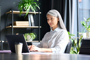 Happy confident older senior Asian businesswoman ceo executive manager sitting at desk working typing on pc laptop computer in contemporary corporation office. Business technologies career concept.