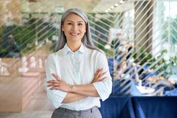 Happy smiling confident middle aged Asian older senior female leader businesswoman standing in modern office workplace looking at camera arms crossed. Business successful executive concept. Portrait. - Powered by Adobe