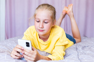 A blonde Caucasian teenage girl lies on the bed with a phone in her hands, plays games, shopping online, blogging. Communicating on social networks, browsing pages. Leisure, rest in a cozy bedroom.