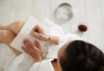 Top view of a brunette woman sitting on the floor and writing in a notebook. Cup with coffee and headphones lie on the floor