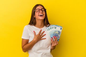 Young caucasian woman holding bills isolated on yellow background laughs out loudly keeping hand on chest.