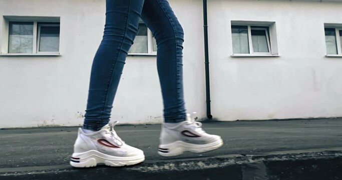 A teenage girl walks along the tarmac along the curb in white crous and jeans. In the background is a white building with windows. 4K.