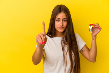 Young caucasian woman holding a home keys isolated on yellow background showing number one with...