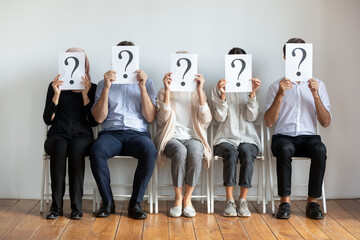 Diverse job candidates hiding faces behind sheets with question marks, sitting on chairs in row,...