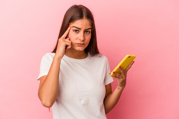 Young caucasian woman holding a mobile phone isolated on pink background pointing temple with finger, thinking, focused on a task.