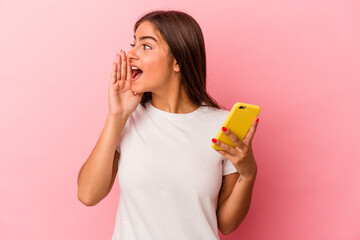 Young caucasian woman holding a mobile phone isolated on pink background shouting and holding palm near opened mouth.