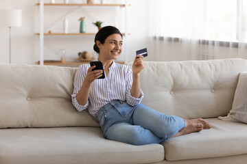 Happy millennial Indian woman relax on sofa in living room shopping online with credit card on smartphone. Smiling young mixed race female pay on internet use secure bank service system on cellphone.