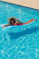 Young smiling African American girl in bikini, wearing straw hat relaxing on inflatable in swimming pool.