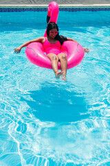 Smiling young African girl in bikini relaxes on inflatable pink flamingo in swimming pool.