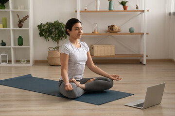Calm young Indian woman sit on mat at home meditate training with online webcam class or lesson on computer. Millennial ethnic female practice yoga with distant web training session on laptop.