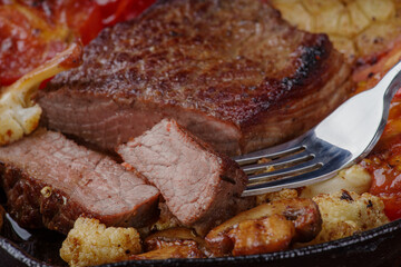 Juicy medium-rare steak in a cast iron skillet with fried vegetables, tomatoes, cauliflower, mushrooms and baked garlic. A juicy piece of marbled beef is cut off and impaled on a fork, taste how delic