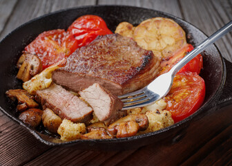 Juicy medium-rare steak in a cast iron skillet with fried vegetables, tomatoes, cauliflower, mushrooms and baked garlic. A juicy piece of marbled beef is cut off and impaled on a fork, taste how delic