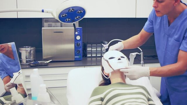 Focused male beautician applying light therapy LED facial mask to woman at esthetic clinic
