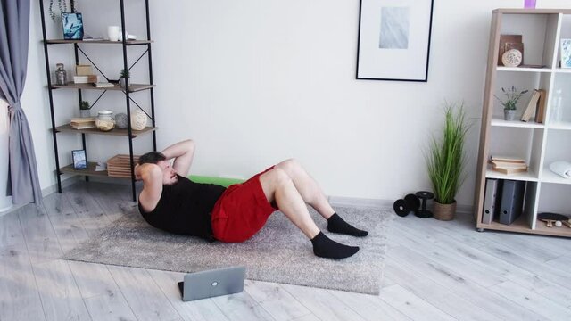 Online fitness group. Exhausted man. Trainer support. Home gym. Chubby guy in shorts t-shirt doing hard exercise trunk curls looking laptop showing like gesture light room interior.