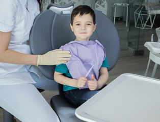 Beautiful child in dentist's chair. 5 years old. Doctor’s hand in a glove on a kid's shoulder. Modern dental equipment. Concept of baby teeth hygiene, caries prevention, treatment without fear.