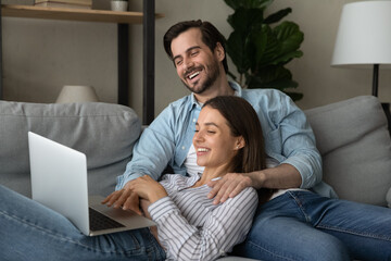 Joyful loving young family couple relaxing together on cozy sofa, watching comedian movie or funny video film online on computer, spending free weekend leisure time using modern technology gadget.
