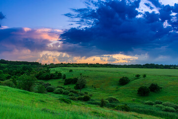 Fototapeta premium Evening twilight over the meadow. Bright dramatic sky and green grass. Rural landscape under a picturesque colorful sky at sunset.