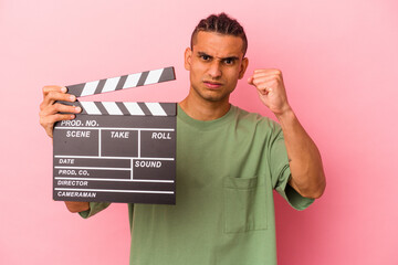 Young venezuelan man holding a clapperboard isolated on pink background showing fist to camera,...