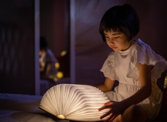 Bella niña asiática leyendo libro luminoso en la cama