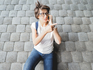 Woman talks by smartphone and points somewhere. Female in jeans and eyeglasses on grey tiled wall background.  Urban lifestyle of millennials. Active communication. Social life.