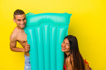 Young latin couple holding an air mattress isolated on yellow background