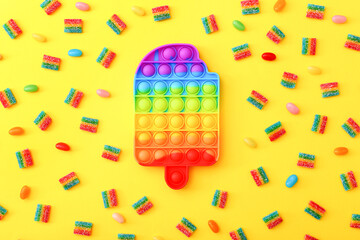 Rainbow Pop it fidget toy next to the colorful marmalade candies on yellow background. - obrazy, fototapety, plakaty