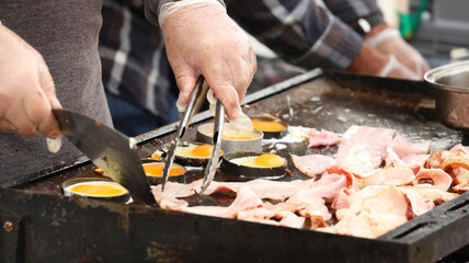 Two hands wearing protective plastic gloves for safe food handling practices cooking bacon on eggs on a sizzling hot community bbq or hot plate grill. - obrazy, fototapety, plakaty