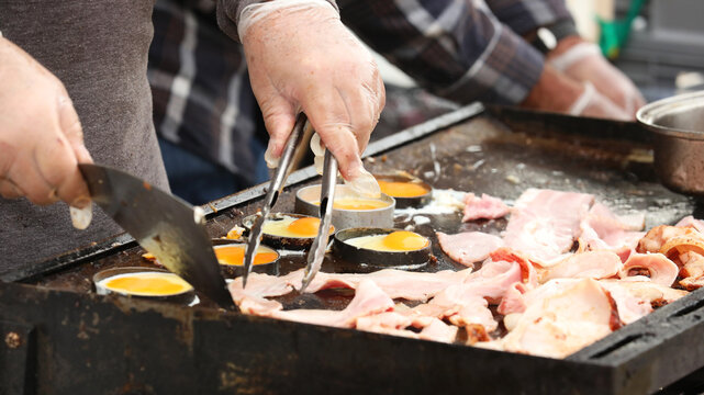 Two hands wearing protective plastic gloves for safe food handling practices cooking bacon on eggs on a sizzling hot community bbq or hot plate grill.