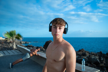 Portrait of a shirtless young attractive male athlete in an open air park,wearing headphones and listening to music to focus.His friend in the background does push-ups.Concept of music during training