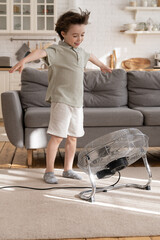Fototapeta Playful small kid alone in living room enjoy fresh cool breeze blowing from big industrial retro ventilator or air conditioner. Cheerful kid has fun indoors with strong cold wind in hot summer season obraz