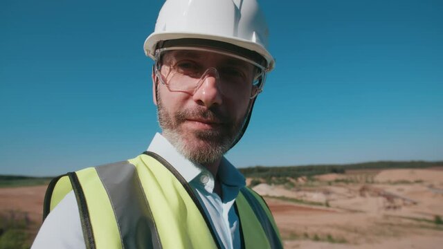 Handheld portrait of positive male sand quarry worker wearing safety uniform goggles hard hat, male caucasian bearded engineer turn to camera and smiling. Sand mining project specialist on sunny day