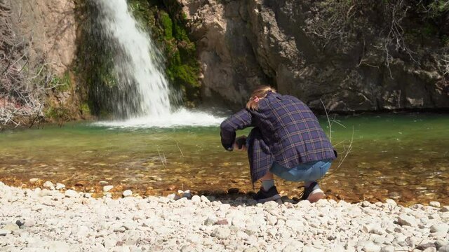 Tourist takes photos of waterfall from low angle while walking in mountain forest, using mobile phone, view from the back. Young female blogger makes content for her travel vlog.