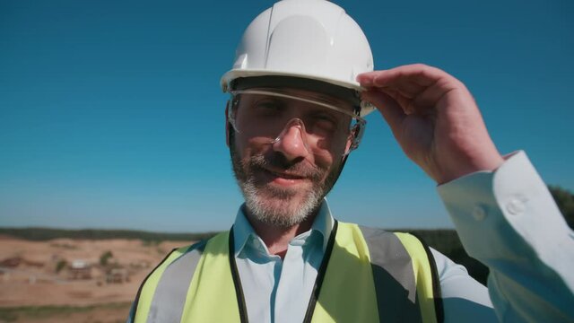 Wide angle portrait positive male sand quarry worker wearing safety uniform goggles and hard hat, male bearded engineer looking at camera and smiling. Sand mining project specialist on warm sunny day