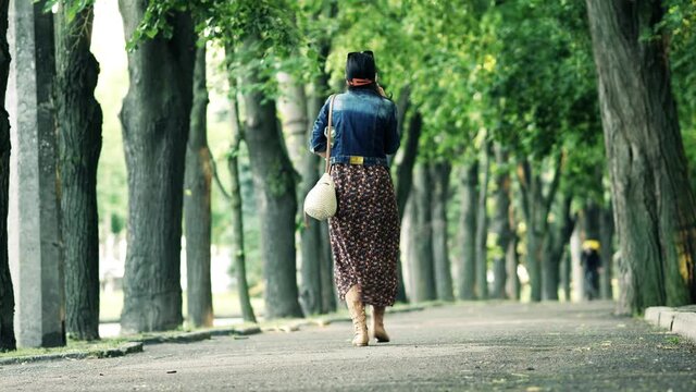 Woman walks along sidewalk on warm summer day. Courier cyclist with yellow backpack   seen in distance. Trees with fresh green foliage grow around. City street during day. Beginning of June.
