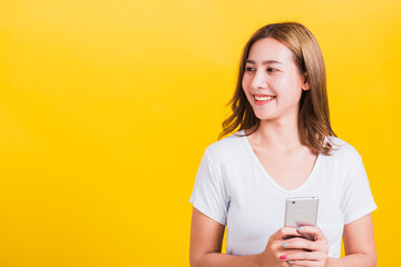 Asian Thai portrait happy beautiful cute young woman smile standing playing game or writing SMS message on smartphone looking to side away space, studio shot isolated on yellow background