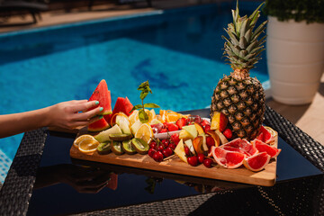 Tropical breakfast by swimming pool, floating breakfast in tropical resort. Healthy breakfast and fruit plate by resort pool. A woman's hand takes a slice of fruit