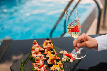 The woman is holding a glass of champagne and a strawberry, and in the background canapés on the table while sitting on the pool. Refreshment on a hot summer day at the pool