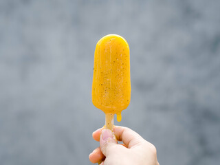 Yellow popsicle in woman hand on grey wall background, melting, summertimes. Female hand holding...