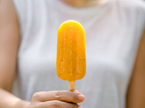 Close Up Yellow Popsicle In Woman’s Hand Wearing Casual White Sleeveless Shirt, Outdoors. Woman Eating Popsicles. Young Female Enjoying Ice Lolly In Summer.