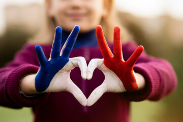 Love and happiness concept. Cute child forming heart gesture with hands outdoors on nature sunset bokeh background. Heart shape of kids hand painted in france flag colors, kids body language
