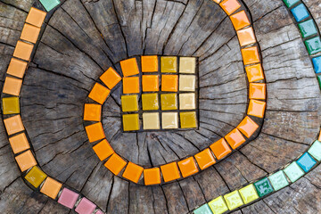 Mosaic tiles on round cut down tree with cracks stump outside. DIY garden furniture, decorated by hand made element small tiles. Mosaic colorful snake path. Abstract natural wooden background