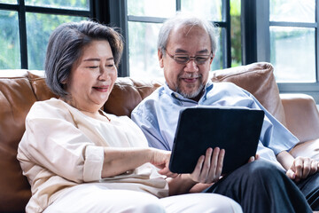 Asian senior couple sit on sofa use tablet read news together in house