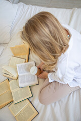 Fototapeta na wymiar Beautiful sensual young woman with blonde hair wearing a white shirt reading books and drinking coffee on the bed in modern home
