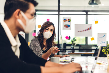 Asian business man and woman using laptop computer working and planning meeting in quarantine for coronavirus wearing protective mask with social distancing while sitting on office desk