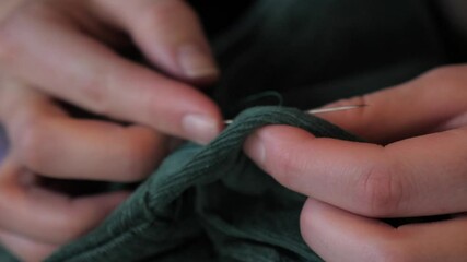 Female hands hand sew green jacket material with thin metal needle and string thread to make alteration and tailoring adjustment, front on macro close up view reaching around with stitching.