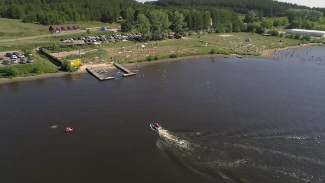 Aerial view of city beach on the outskirts of a small provincial town. People lie on the sand, swim in a pond, relax, dive, ride a jet ski. Near forest and parking with many cars. Summer sunny day
