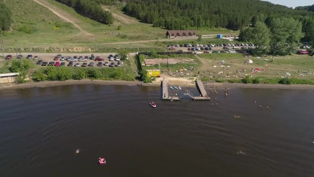 Aerial view of city beach on the outskirts of a small provincial town. People lie on the grass, swim in a pond, relax, dive, ride a jet ski. Near forest and parking with many cars. Summer sunny day
