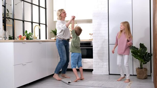 Excited mom or babysiter and two kids boy and girl jumping, dancing laughing in modern scandinavian house kitchen. Happy family mother daughter son having fun enjoy playing singing together at home.