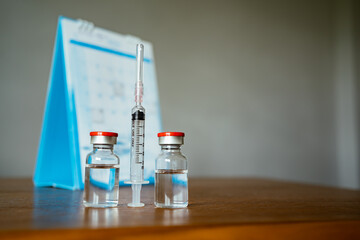 2 vials of vaccine, syringe and a calendar in the background