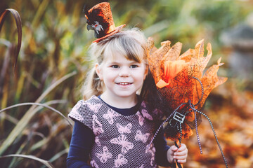 Portrait of little toddler girl dressed as a witch celebrates Halloween. Happy child outdoors, with...